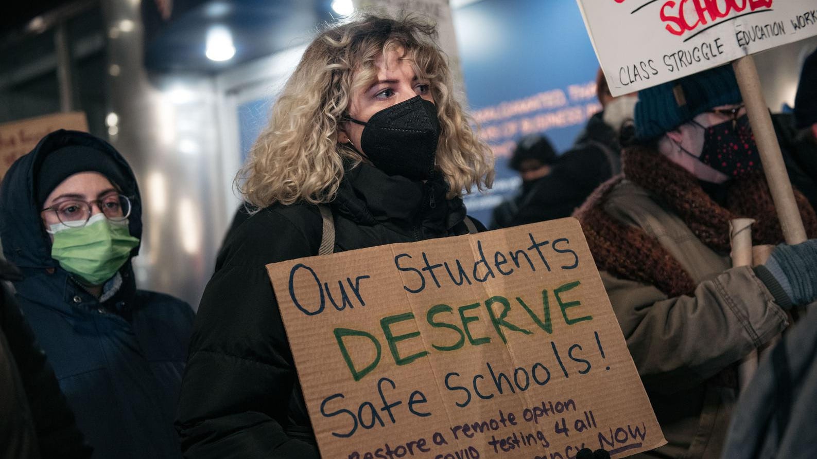 A woman wears a black mask during a protest, the sign says, "our students deserve safe schools"