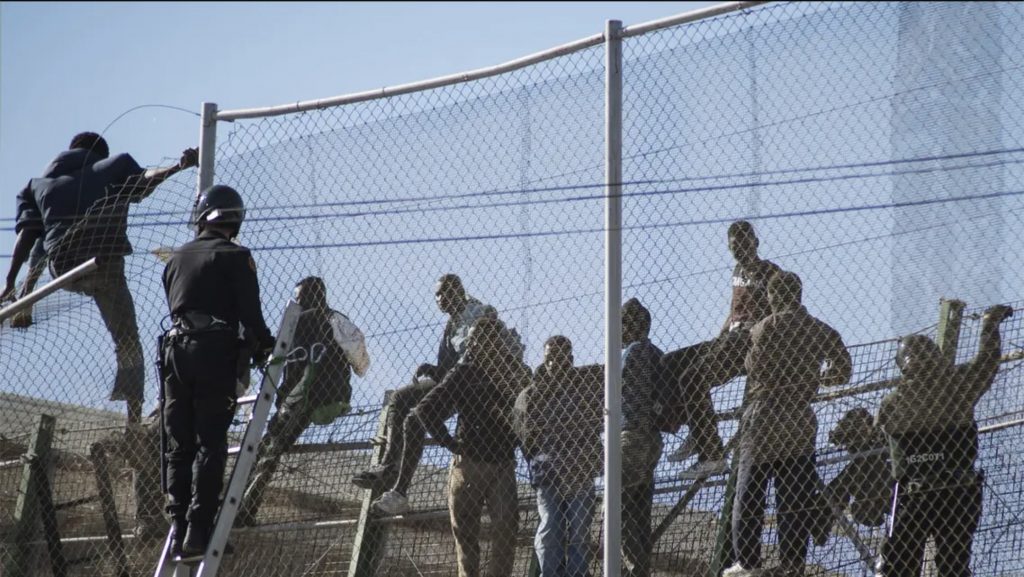 Police scale the fence from the left, approaching a group of migrants that are sitting on the fence.