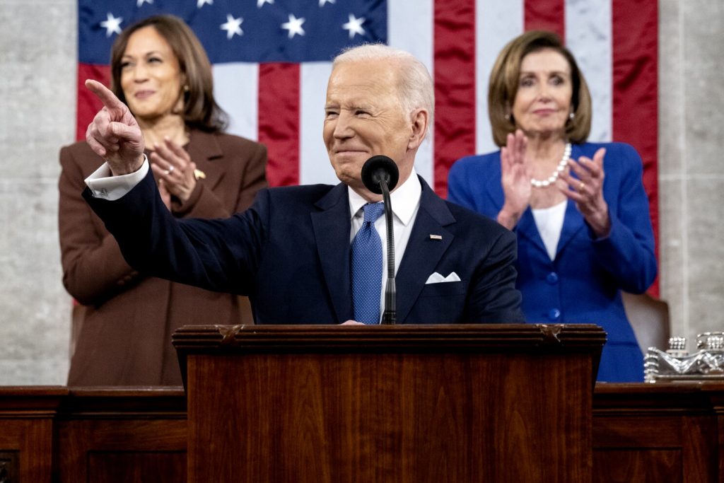 President Biden stands at a podium at his 2022 State of the Union Address. Kamala Harris and Nancy Pelosi stand behind him.