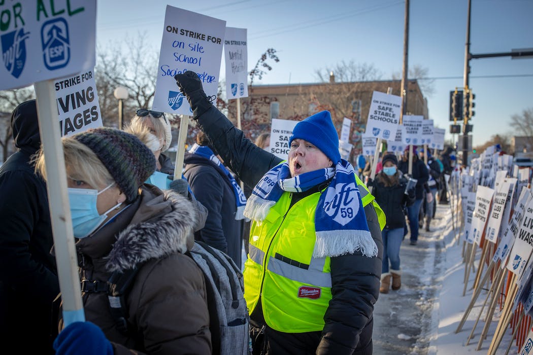 Surrounded by striking workers holding up picket signs, a person in the center is wearing a bright blue beanie and MFT scarf and a bright yellow vest on top of a black undershirt and black pants.