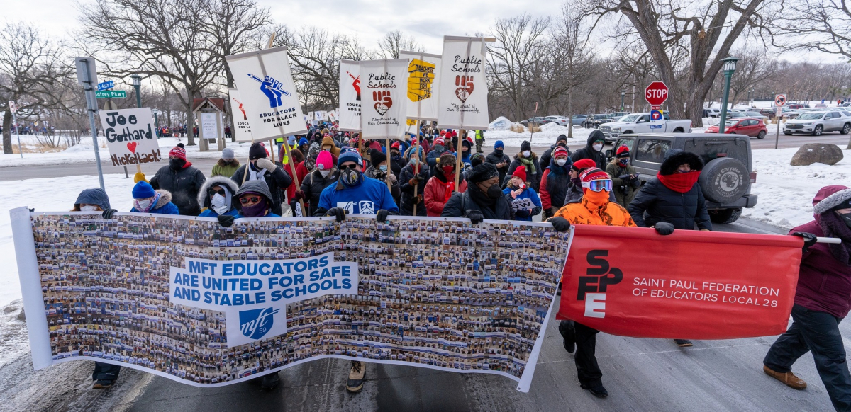 Minnesota teachers hold two banners, one saying "MFT Educators are United for Safe and Stable Schools." in blue text. The other banner being held up by teachers is red and says, "Saint Paul Federation of Teachers Local 28," in white text on the right with the local symbol on the left.