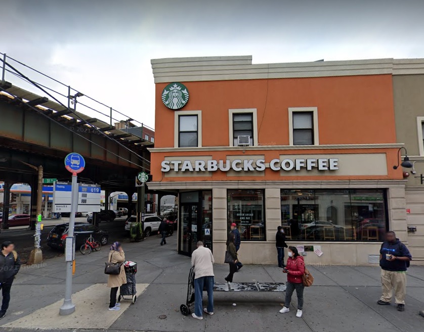 On the left is a blue bus stop for the Q19 bus as well as the subway tracks. On the right is a brown building with the Starbucks logo in the upper left corner, the starbucks coffee sign in white lettering in the middle of the building. Some people gather in front of the store.