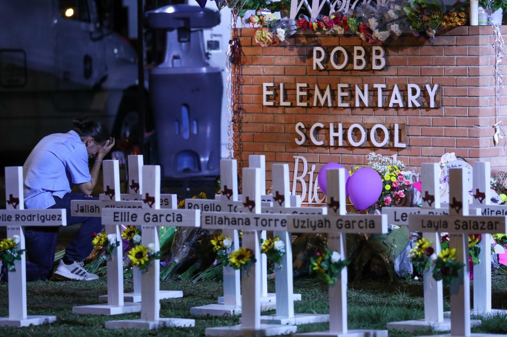 A collection of white crosses with the murdered children's names on them in front of a Robb Elementary School sign. A kneeling adult cries off to the side