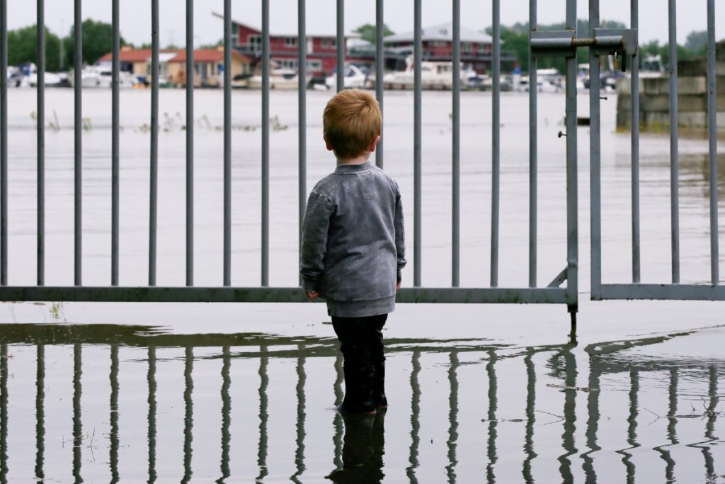 A young child stands in ankle deep water in front of a metal fence.