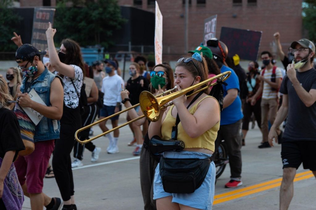 A protest, people are milling around. In the center of the frame is a woman playing the trombone.