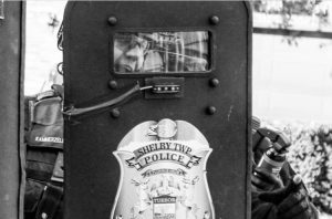 Police office crouches behind a riot shield which reads "Shelby Township Police"