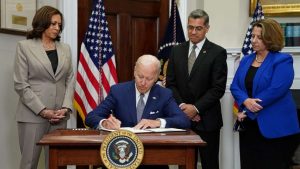 Vice President Kamala Harris stands on the left, Biden in the center signing a piece of paper at a desk with the US flag behind them and between them, and Health and Human Services Secretary Xavier Becerra and Deputy Attorney General Lisa Monaco stand at his right side.