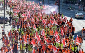 Dock workers march holding red flags and wearing yellow and orange construction vests.