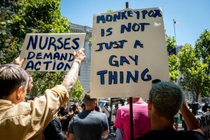 On the left a white sign with blue text reads "nurses demand action!" in all caps and on the right a sign reads in blue capitalized text "monkeypox is not just a gay thing"