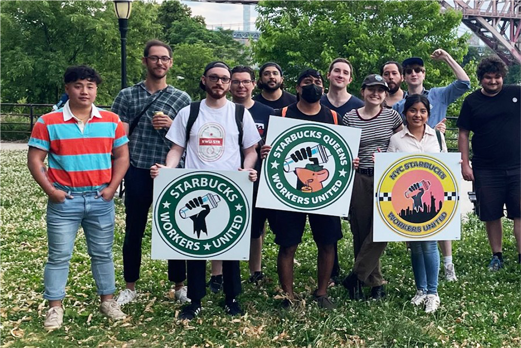 Starbucks workers from Queens, New York stand in a park and hold Starbucks Workers United signs.