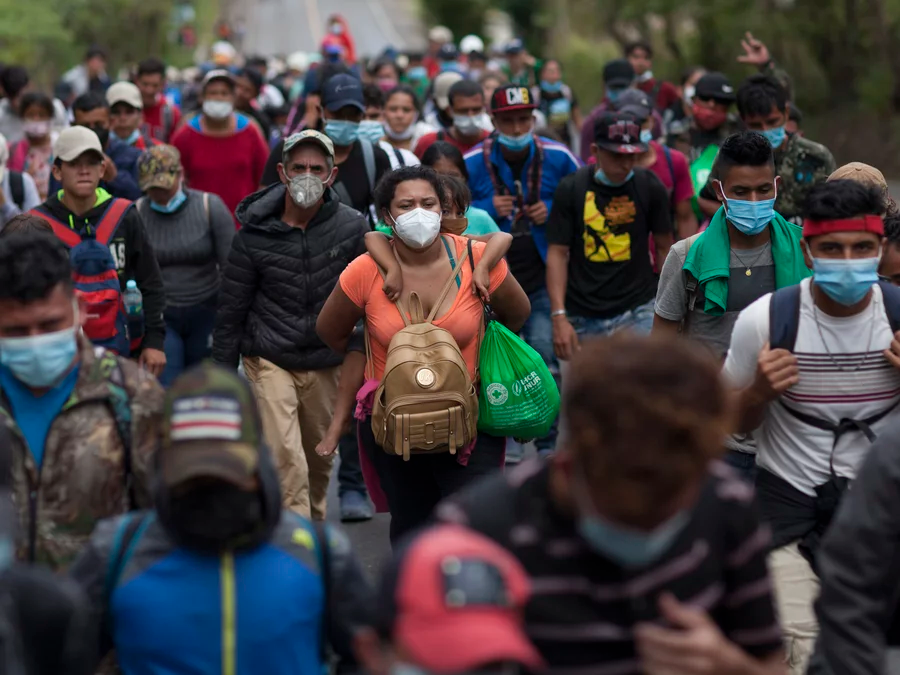 Image of large crowd with people wearing surgical masks. Image is centered and focused on a woman carrying a child on her back.