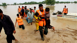 A male rescue worker rescues two children from flood waters, with two rescue boats and other people in the background.