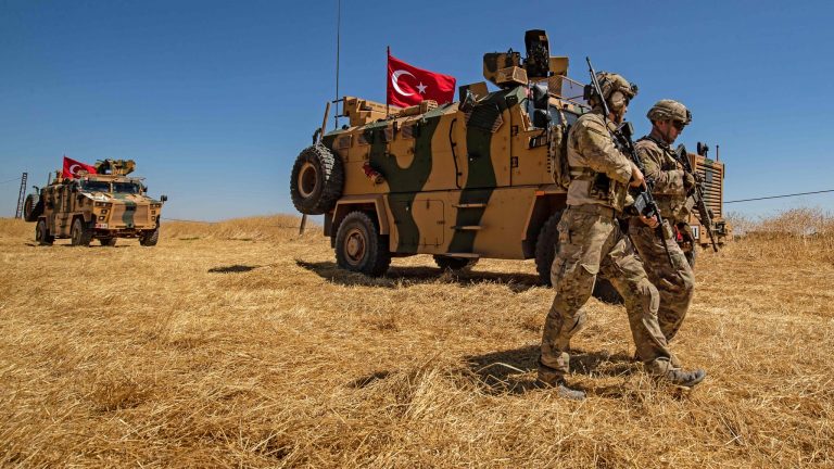 Two Turkish military members stand in front of a tank with the Turkish flag in the foreground and another tank with military members is visible in the background.