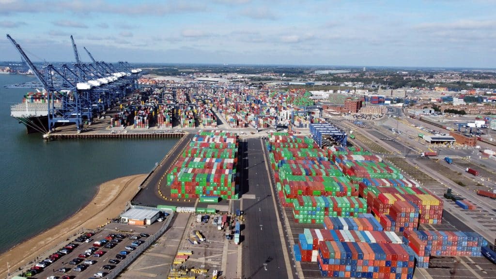 Photograph of a port, with water on the left, shipping containers in green and red in the left foreground, a road in the center, shipping containers in red and green in the background, and shipping containers in red, blue, and white in the foreground.
