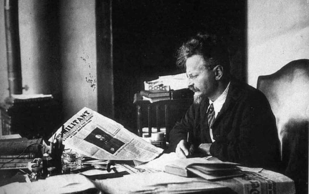 A black and white photograph of Leon Trotsky seated at a desk reading a newspaper titled "The Militant".