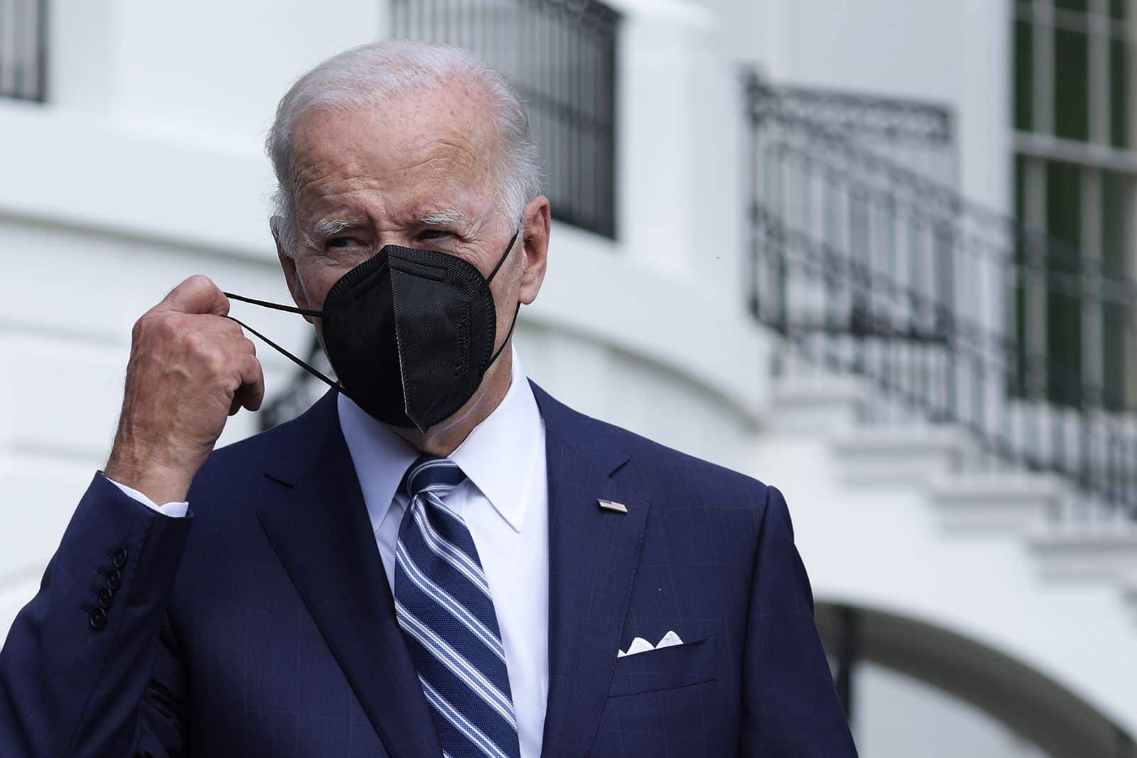 US President Joe Biden stands in a suit wearing a mask, but is taking off one side of it.