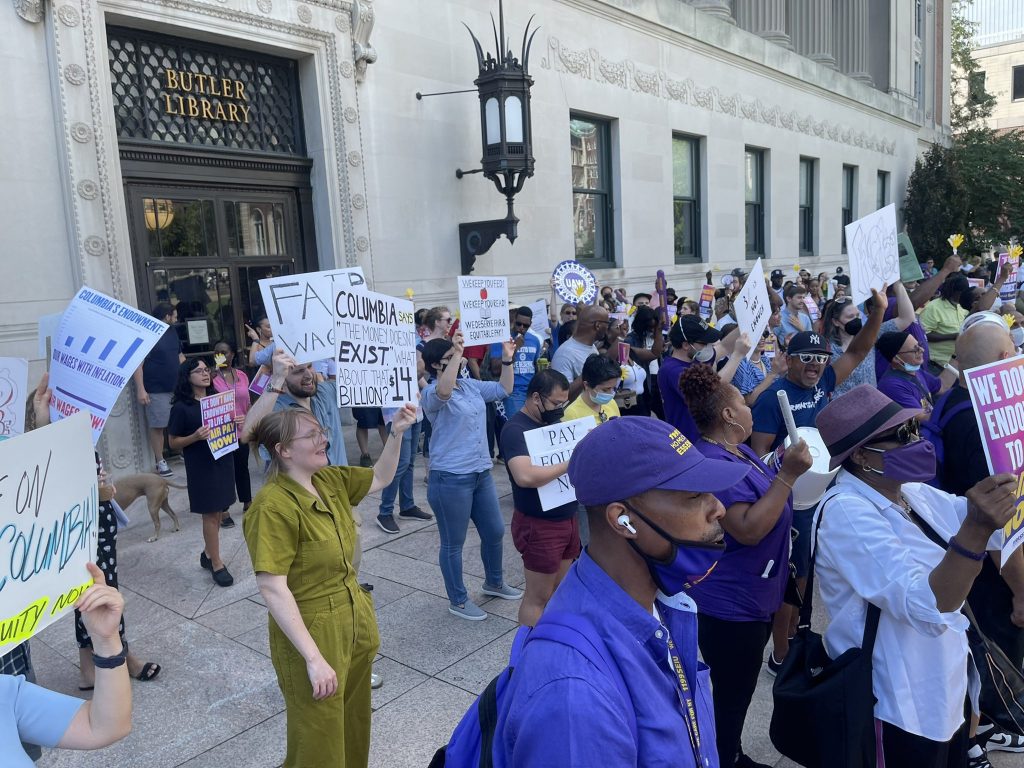 A crowd of about 100 workers and their allies, many of them dressed in SEIU purple, gather with signs in front of Columbia's Butler Library to demand bigger raises