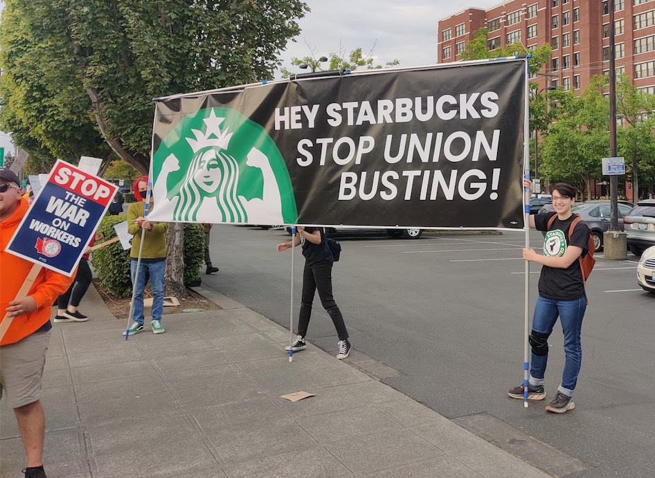 Protestors on a sidewalk holding a vinyl banner that says "Hey Starbucks, stop union busting!" In the foreground is another protestor holding a picket sign that says "stop the war on workers!"