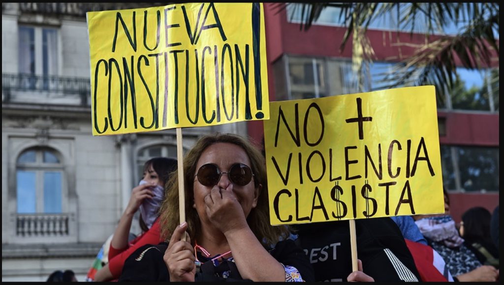 Woman holds a yellow sign that says "Nueva constitucion" in black text and another person is holding a yellow sign behind her that reads "No violencia clasista"