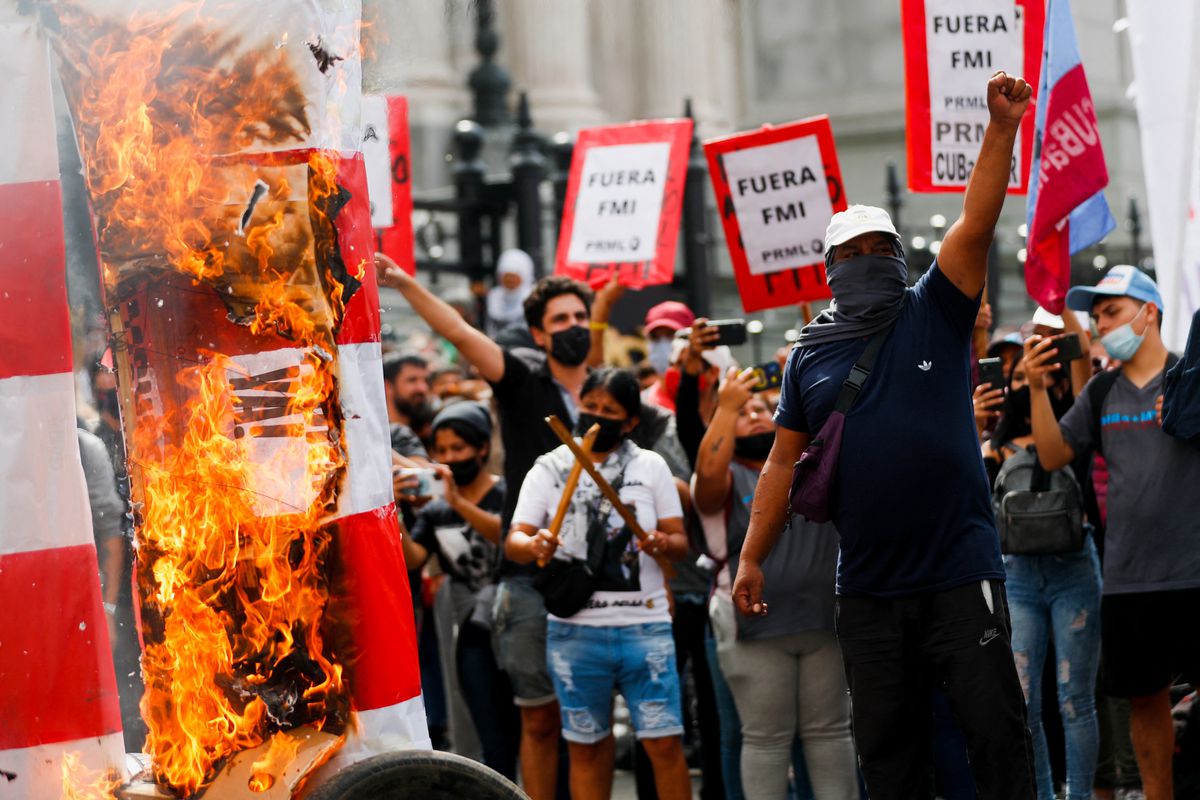 On the far left, a sign is on fire. Protestors stand and hold signs with black text on white backgrounds mounted on red posters that read "Fuera FMI!" or "Out with the IMF!"