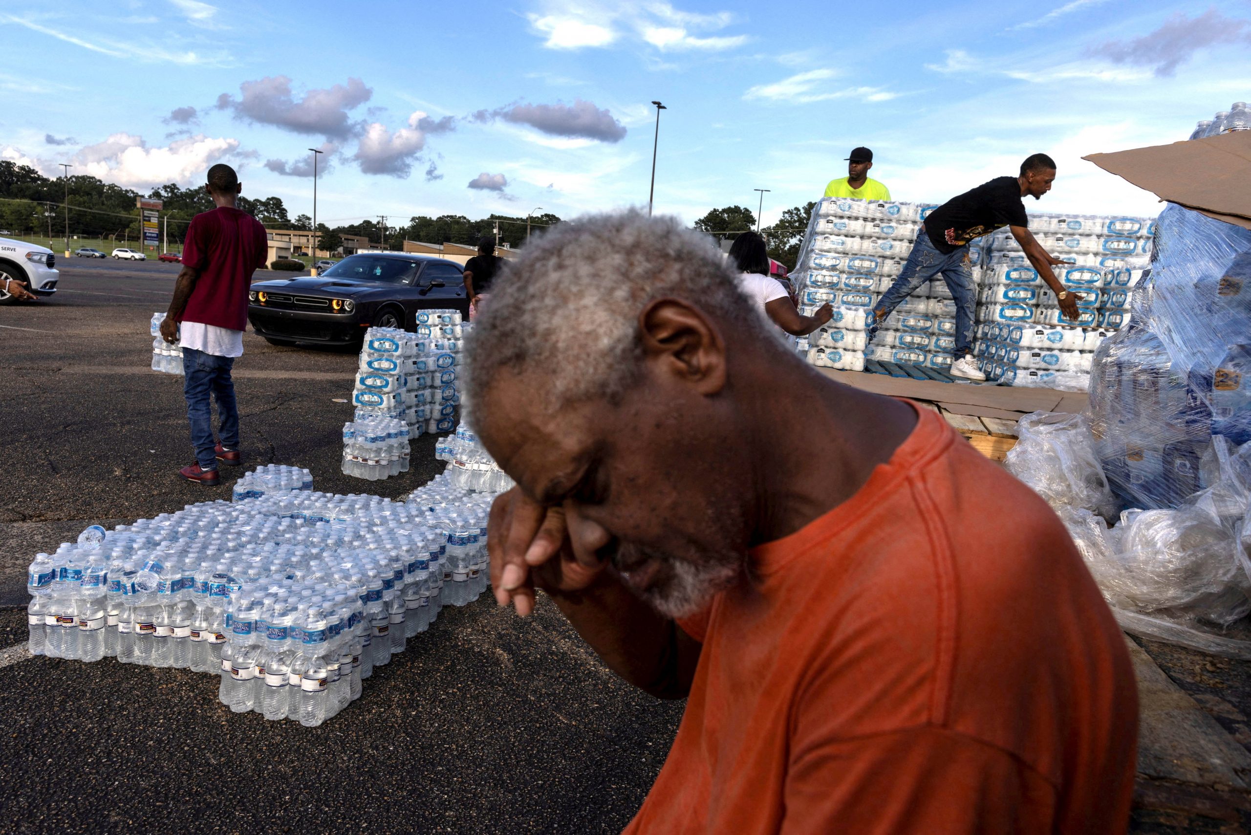 In the center foreground, a Black person with gray close shaved hair wearing an orange t shirt wipes their face with their hand. In the background are pallets of water bottles and people are moving them.