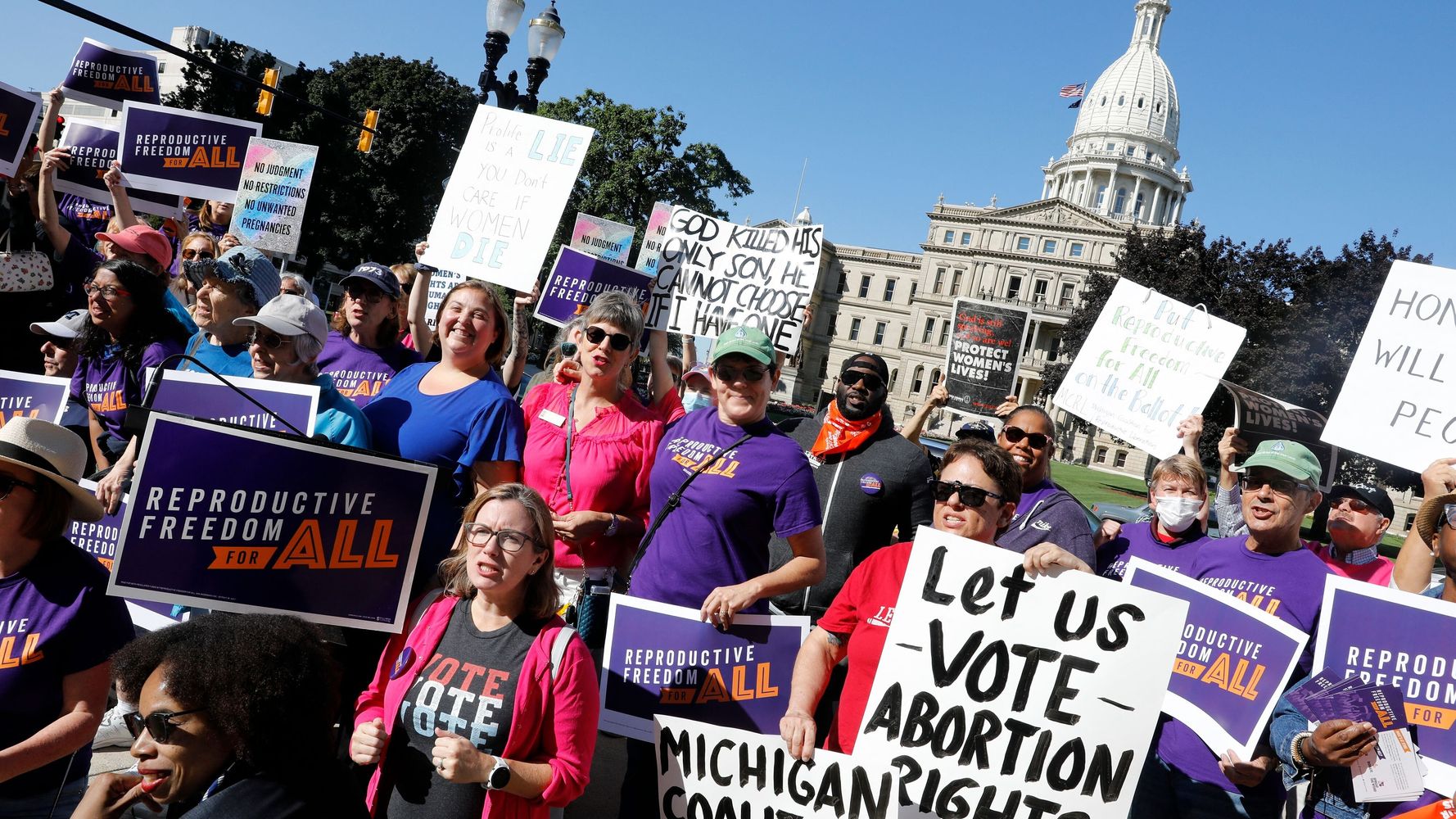 A group of protesters is standing in front of a white government building wearing purple shirts that say "reproductive freedom" in white text and "for all" in orange text. A protester in the front right holds a sign that says "Let us VOTE abortion rights," and many of the protesters are holding purple signs that also say "reproductive freedom" in white text and "for all" in orange text.