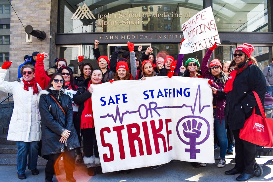 Several nurses, all wearing red, stand together in front of Mount Sinai hospital with raised fists and signs reading "Safe Staffing or Strike"