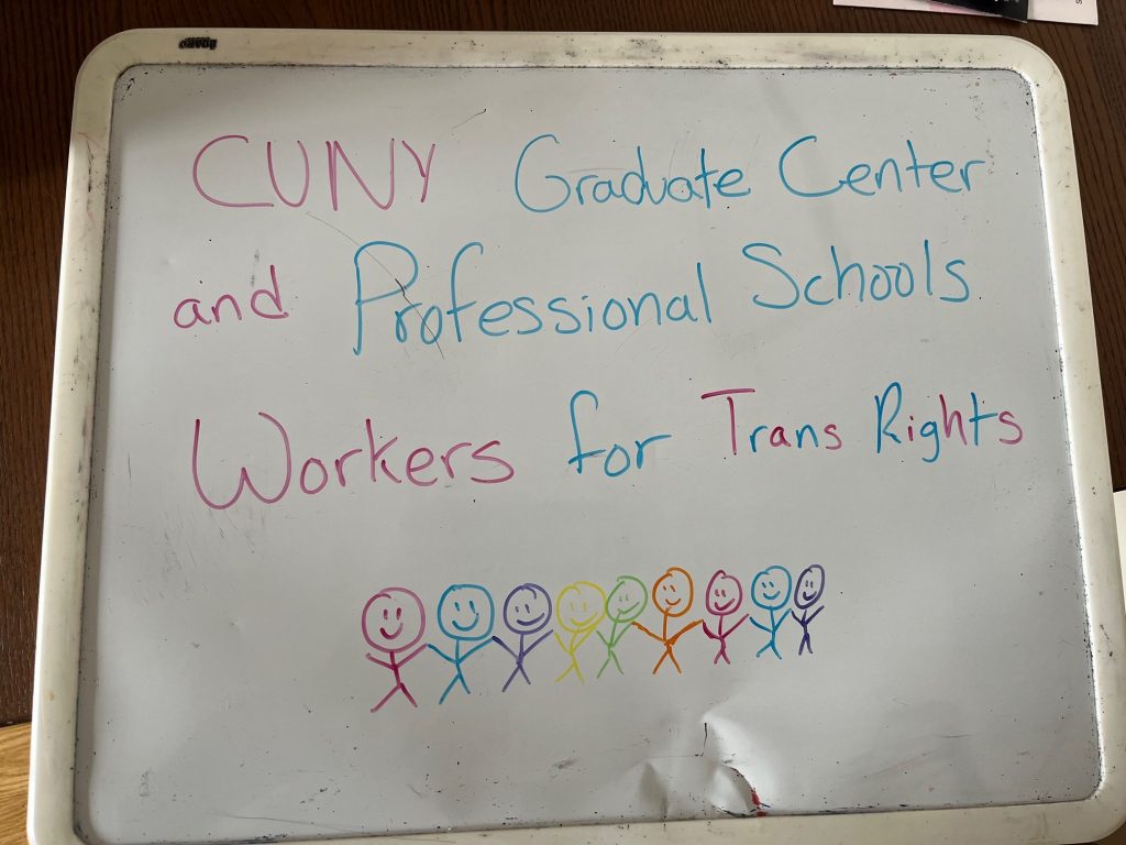 A sign drawn on a small whiteboard, in trans pride colors. Text: "CUNY Graduate Center and Professional Schools Workers for Trans Rights" Beneath the text is a chain of 9 smiling stick figures in different colors, all holding hands
