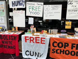 Three tables full of food, with signs hung above them. One says "The People's Pantry: FREE FOOD." Banners hung from the tables say "Free CUNY" and "Cop Free School Zone"