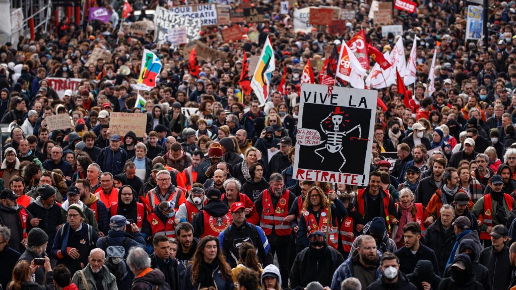 A group of protesters, in the front of whom are a line of protesters wearing red vests. In the front right corner, a white sign reds "vive la retraite," with a skeleton wearing a red hat in the middle of the sign on a black background with a text bubble on its left that reads, "oiv a bosse, c'est pas pour en crever!"