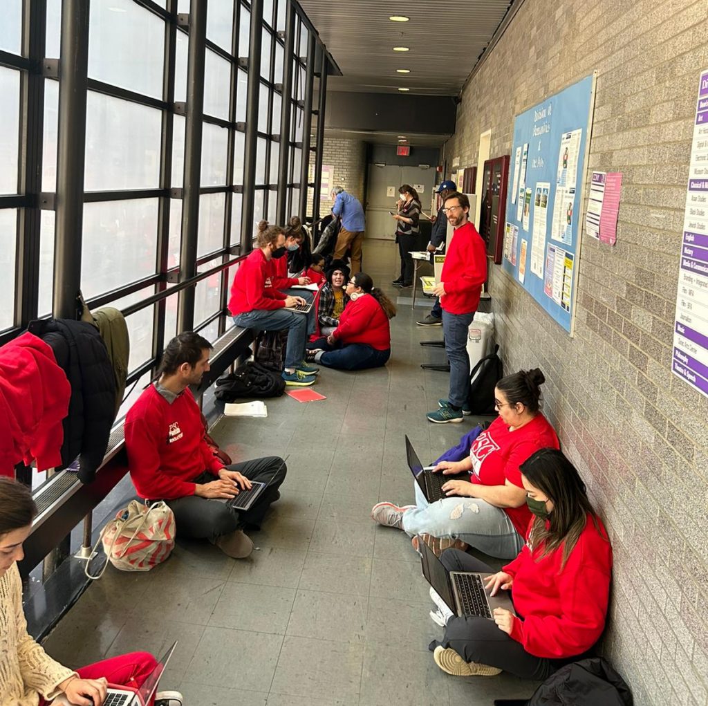 Twelve or so faculty members wearing red union sweatshirts standing and sitting with laptops in a hallway