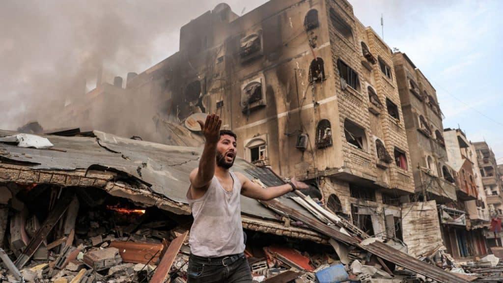 A Palestinian man in a white tanktop stands in front of a burnt-out apartment building in Gaza during the Israeli assault in October 2023.