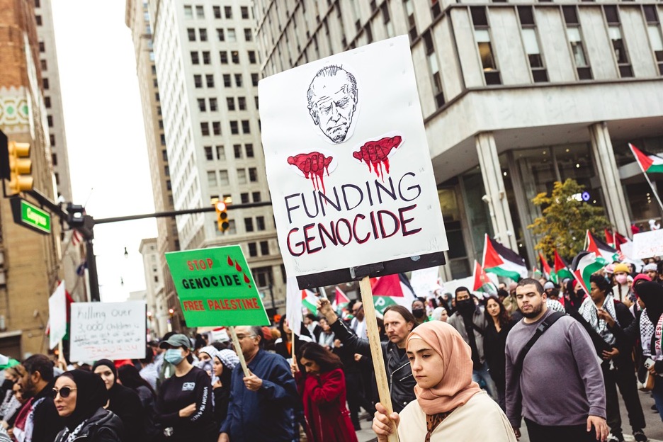 A protester at a pro-Palestine protest in Detroit in October 2023, wearing a hijab, holding a sign that reads "Funding Genocide" with a picture of Joe Biden. 