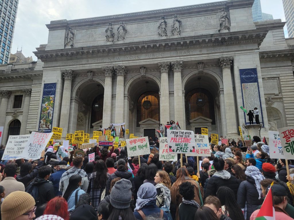 A crowd of activists holding signs with slogans like "Palestine Will Be Free" and "Defend Gaza Against Israeli Terror" gather in front of the NY Public Library
