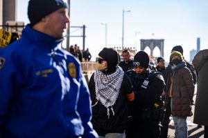 A NYPD officer arrests a masked pro-Palestine protestors wearing a kaffiyeh while other cops and masked protesters look on.