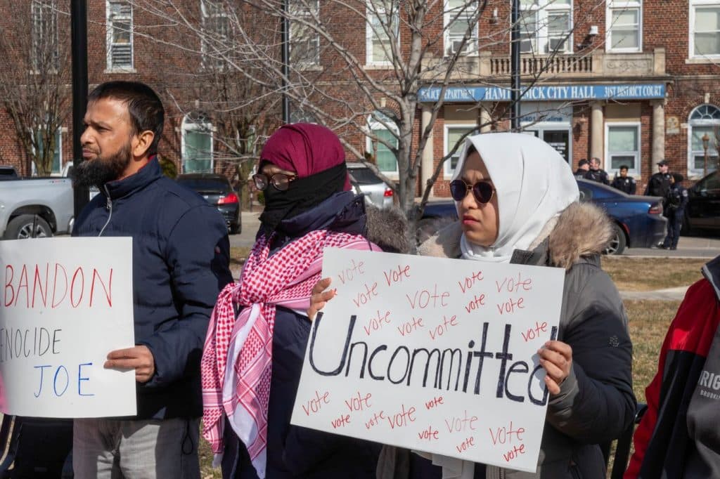 Three activists stand together while one activist holds up an "uncommitted" sign with the word "vote" written on it many times. In the left side of the picture another activist holds a sign that says "abandon Genocide Joe"
