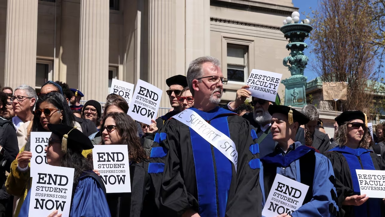 A group of Columbia University faculty dressed in regalia hold signs that say "end student suspensions now"